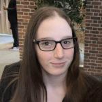 Lexi Hoare head shot photo, a young white woman with long straight brown hair and black eyeglasses wearing a black blazer