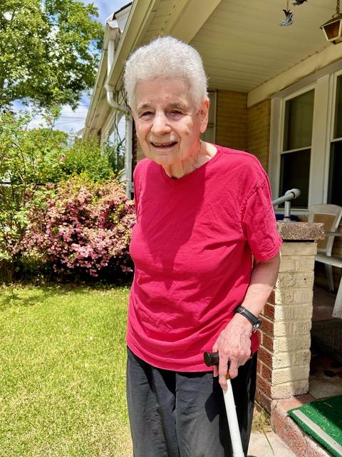 Joyce Driben, an older woman with white hair, a red shirt and black slacks on, stands with a walking cane in her backyard garden.