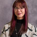 Taylory Akers, a white woman with long reddish brown hair and eyeglasses, wearing a black turtleneck with a white striped blazer over it.